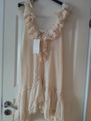 Beige volangklänning från Zara - Supersöt beige klänning från Zara med volanger längs urringningen och nederkanten. Ärmlös modell med v-ringning och romantisk känsla. Perfekt för sommarens alla tillfällen. Tillverkad i ett lätt och luftigt material.