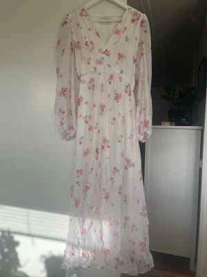 Vit blommig långklänning Neo Noir - Romantisk långklänning från Neo Noir i vitt med rosa blommigt mönster. Klänningen har v-ringning, långa ärmar och volangdetalj nertill. Perfekt för sommarens alla tillfällen. Orginalpris ligger på 1200kr 