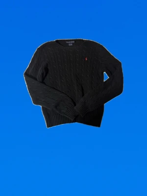 Svart kabelstickad tröja Ralph Lauren - Kabelstickad svart tröja från Ralph Lauren med rund halsringning och diskret röd logga broderad på bröstet. Tröjan har långa ärmar och ribbade muddar vid ärmslut och nederkant. Perfekt för en stilren och klassisk look.