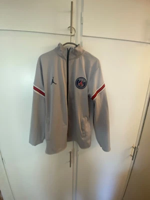 Grå PSG x Jordan ziptröja - Snygg grå ziptröja från Jordan i samarbete med PSG. Tröjan har dragkedja, hög krage och PSG-logga på bröstet samt Jordan-logga. Röda och blå ränder på ärmarna ger en sportig look. Perfekt för fotbollsfans och streetstyle.