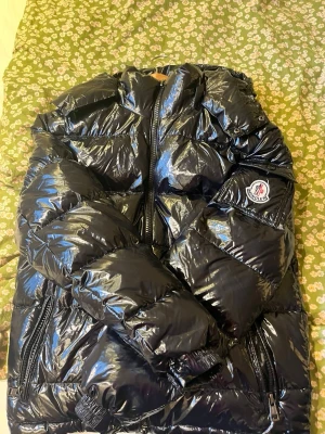 Svart glansig dunjacka från Moncler - Säljer en svart, glansig dunjacka från Moncler med klassisk logotyp på ärmen. Jackan har dragkedja framtill, fickor med dragkedja och en puffig, quiltad design som ger en riktigt trendig look. Perfekt för kalla vinterdagar och riktigt snygg till streetwear.