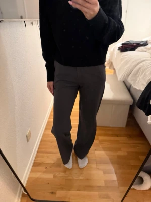 Mörkgrå kostymbyxor med rak passform - Snygga mörkgrå kostymbyxor med rak passform och hög midja. Materialet ser stretchigt och bekvämt ut, perfekt för en avslappnad men ändå stilren look. Inga synliga fickor eller detaljer, vilket ger en clean och minimalistisk stil.