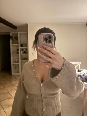 Beige kofta med pärlknappar - Slutsåld beige kofta från Gina med djup v-ringning och pärlknappar framtill. Koftan har breda, långa ärmar och ribbad midja som ger en snygg passform. Perfekt att styla med jeans eller kjol för en cool look. Tröjan är lite sönder i midjan men går att fixa enkelt💞