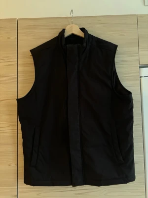 Svart dunväst med hög krage - Svart dunväst med hög krage och dold dragkedja framtill. Västen har två sidofickor och en rak passform. Strl: M. Använd en del i tidigare dagar men fortsatt i bra skick.