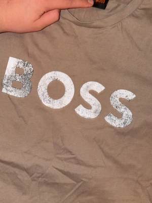 Beige BOSS t-shirt med tryck - Snygg beige t-shirt från BOSS med stort vitt logotryck över bröstet. T-shirten har klassisk rund hals och korta ärmar. Perfekt för dig som gillar stilrena plagg med tydlig branding.