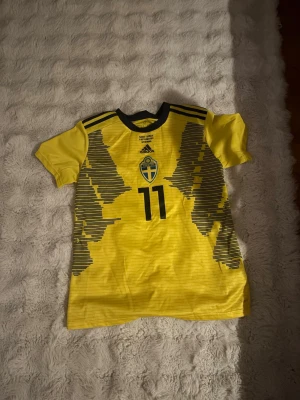 Sverige matchtröja Blackstenius #11 - Gul matchtröja från Adidas för svenska landslaget med Blackstenius och nummer 11 på ryggen. Tröjan har svarta detaljer, landslagets emblem på bröstet och korta ärmar. Perfekt för fotbollsfans som vill stötta Sverige på matchdagar.