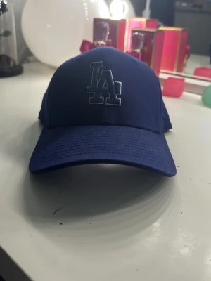 Blå LA keps från New Era - Snygg blå keps med LA-logga framtill från New Era. Klassisk böjd skärm och justerbar baksida. Perfekt för dig som gillar sportiga accessoarer och vill ha en cool look. Materialet är bomull 