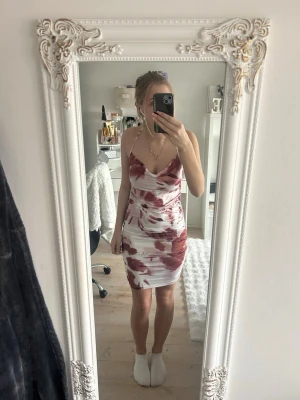 Kort klänning med brunröd mönster  - Snygg kort klänning med smala axelband och v-ringad front. Klänningen har ett abstrakt brunrött mönster på vit botten och korsade band i ryggen. Materialet känns lätt och passar perfekt för varma dagar. Stilren och trendig design.