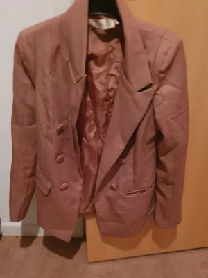 Kavaj - Snygg brun kavaj med dubbelknäppning och breda slag. Kavajen har två fickor framtill med lock och matchande knappar. Perfekt för dig som vill ha en klassisk och stilren look. Materialet känns mjukt och har en lätt glans.