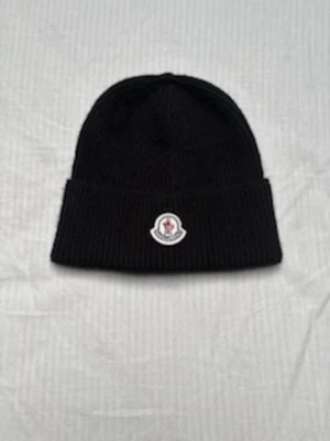 Svart Moncler mössa i ribbstickad ull - Svart ribbstickad mössa från Moncler med uppvikt kant och klassisk logotyp framtill. Tillverkad i mjuk ull som håller dig varm och snygg under kalla dagar. Enkel och stilren design som passar till det mesta.