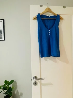 Blå stickad topp med knappar - Snygg blå stickad topp med v-ringning och tre små knappar framtill. Tillverkad i ett lätt, strukturerat tyg som ger en avslappnad känsla. Perfekt att matcha med jeans eller kjol för en trendig look.