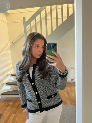 Svartvit kofta med guldknappar - Säljer en snygg svartvit kofta med guldknappar och svarta detaljer vid fickor, ärmslut och nederkant. Koftan har ett klassiskt mönster och två fickor framtill. Perfekt för att lyfta din outfit.