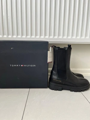 Tommy Hilfiger leather chelseaboots mid calf boots - Extremt fina Tommy Hilfiger chelseaboots i läder material som är helt nya! Prislappen och skokartongen finns kvar, skorna ligger i kartongen och är redo att fraktas! Svart färg med diskret Tommy Hilfiger logga i guld färg  Storlek 39 Köpt ifrån Zalando, nypris 2000kr Skicka meddelande vid några funderingar Kolla gärna även in mina andra annonser