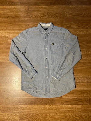 Blå skjorta från Lyle & Scott - Säljer en klassisk blå skjorta från Lyle & Scott med button-down krage och bröstficka med broderad logotyp. Skjortan har långa ärmar och knappar framtill. Perfekt för dig som gillar stilrena plagg med diskreta detaljer.
