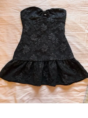Svart axelbandslös blommig klänning - Säljer en svart axelbandslös kortklänning med blommigt mönster och volang nedtill. Klänningen har en söt knutdetalj framtill och är perfekt för dig som gillar en feminin och trendig stil. Köpt på en marknad i Spanien. 