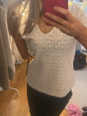 Vit spetsblus med v-ringning - Söt vit blus med v-ringning och halvlånga ärmar. Blusen har ett spetsliknande hålmönster och är lätt figursydd för en feminin look. Perfekt till jeans eller kjol för en fräsch stil.