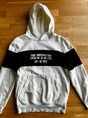 Vit hoodie med svart panel och tryck - Säljer en stilren hoodie i vitt med bred svart panel över bröstet och armarna. Hoodien har stor huva och magficka framtill. Perfekt för dig som gillar streetwear vibes. Den är i mycket fint skick. 