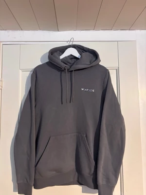 Grå hoodie från McKenzie - Snygg grå hoodie från McKenzie med vit logga på bröstet och stort tryck på ryggen. Klassisk känguruficka framtill och justerbar huva med dragsko. Tillverkad i mjukt bomullsmaterial som känns skönt mot huden. Perfekt för en avslappnad streetwear-look.