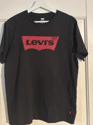 Svart Levi's t-shirt med röd logga - Klassisk svart t-shirt från Levi's med den ikoniska röda loggan tryckt på bröstet. T-shirten har rund halsringning och korta ärmar. Tillverkad i mjuk bomull för en skön och avslappnad stil.
