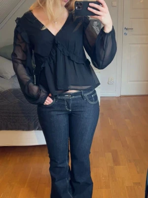 Svart blus - Snygg svart blus i transparent material med volangdetaljer framtill och v-ringning. Blusen har långa ärmar i tunn mesh och avslappnad passform.