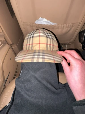 Burberry beige keps med rutmönster - Säljer en beige keps från Burberry med klassiskt rutmönster i brunt, svart och rött. Kepsen har justerbar snapback och en svart patch med texten 'BURBERRY LONDON ENGLAND' på baksidan. Perfekt för dig som vill ha en ikonisk accessoar.