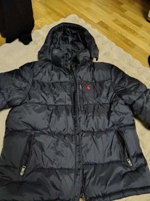 Svart pufferjacka från Polo Ralph Lauren - Snygg svart pufferjacka från Polo Ralph Lauren med huva och dragkedja framtill. Jackan har två framfickor med dragkedja och den klassiska röda loggan på bröstet. Perfekt för kalla vinterdagar och riktigt skön att bära.