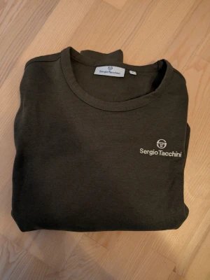 Mörkgrön sweatshirt från Sergio Tacchini - Mörkgrön sweatshirt från Sergio Tacchini med rund hals och diskret logotyp på bröstet. Tröjan har långa ärmar och ribbade muddar vid ärmslut och nederkant. Storlek M, väldigt fint skick.