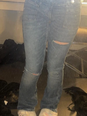 Blå slitna jeans med raka ben från Gina - Säljer ett par blå jeans med raka ben och slitningar på knäna. Jeansen har stora framfickor, hög midja och dubbla knappar i linningen. Materialet är klassisk denim i bomull och de har en avslappnad passform.