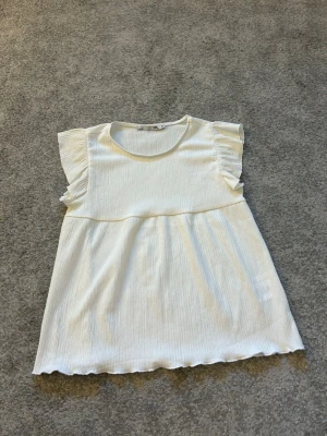 Vit ribbad topp med volangärm 157 - Söt vit topp från 157 med ribbad struktur och volangdetaljer på ärmarna. Modellen är lös och har rund halsringning samt lätt vågig kant nertill. Perfekt för en avslappnad och trendig look.