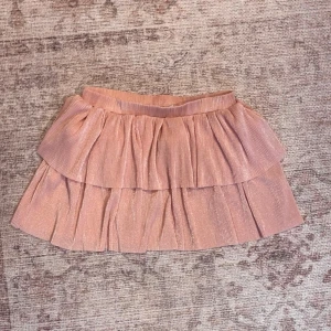 Glittrig rosa volangkjol - Supersöt kortkjol i rosa med glittrigt tyg och dubbla volanger. Kjolen har ett elastiskt midjeband och ett skirt, plisserat material som ger extra volym och en lekfull look. Perfekt för dig som vill sticka ut lite extra!