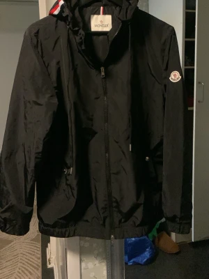 Svart Moncler vindjacka med huva - Svart vindjacka från Moncler med huva och dragkedja framtill. Jackan har Moncler-logga på vänster ärm och diskreta fickor med dragkedja. Materialet är lätt och vattenavvisande, perfekt för blåsiga dagar. Huva med rödvita detaljer på kanten.