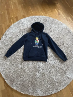  Polo Ralph Lauren Hoodie  - Mörkblå hoodie från Ralph Lauren med den ikoniska Polo Bear-figuren på bröstet. Hoodien har huva med dragsko och en stor magficka framtill. Tillverkad i mjukt material som känns skönt mot huden. Perfekt för en avslappnad och stilren look.