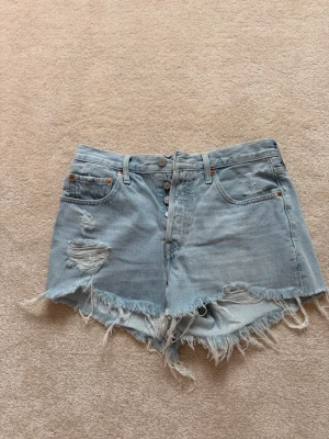 Levi's ljusblå jeansshorts med fransar - Säljer ett par ljusblå jeansshorts från Levi's med fransiga kanter. Shortsen har slitna detaljer framtill, klassiska fem fickor och knappgylf. Perfekta för sommaren och ger en avslappnad vibe. Köpt för ca 1200 kr
