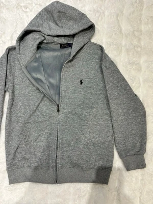 Grå hoodie från Polo Ralph Lauren - Snygg grå hoodie från Polo Ralph Lauren med dragkedja framtill och klassisk huva. Tröjan har det ikoniska svarta Polo-logotrycket på bröstet och ribbade muddar vid ärmar och nederkant. Tillverkad i mjukt bomullsmaterial för extra komfort.