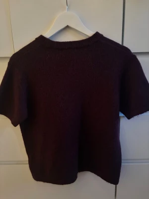 Vinröd stickad t-shirt Gina Tricot - Kortärmad stickad tröja i vinröd från Gina Tricot. Tröjan har rund halsringning och är gjord i ett mjukt, lite luddigt material. Perfekt för lager-på-lager och en snygg basic i garderoben.