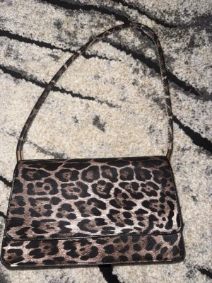 Leopardmönstrad axelremsväska - Snygg handväska med leopardmönster i brunt och svart. Väskan har en rektangulär form, axelrem i samma mönster och guldiga detaljer vid remfästena. Invändigt finns flera fack för enkel organisering. Perfekt för dig som vill sticka ut med ett djurmönster.