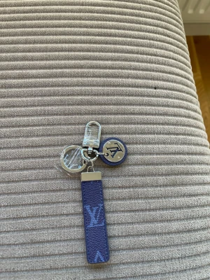  Vuitton blå nyckelring - Säljer en snygg blå nyckelring från  Vuitton med silverfärgade metalldetaljer och LV-logga. Nyckelringen har både en rund bricka och ett avlångt läderband med monogram. Perfekt att hänga på väskan eller nycklarna.
