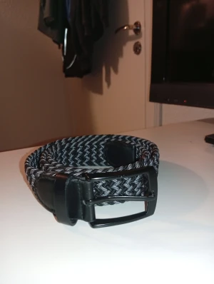 Svart och grått flätat bälte - Snyggt flätat bälte i svart och grått med klassiskt metallspänne. Bältet har detaljer i konstläder vid spännet och ändarna, vilket ger en stilren look. Perfekt accessoar för att lyfta din outfit.