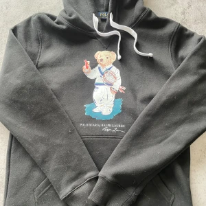 Svart hoodie Polo Bear Ralph Lauren - Svart hoodie från Ralph Lauren med den ikoniska Polo Bear-trycket på bröstet. Hoodien har vit dragsko i huvan och känguruficka framtill. Tillverkad i mjukt material, perfekt för en avslappnad och stilren look. Modellen är 181cm lång. PRIS GÅR ATT DISKUTERA!!! 
