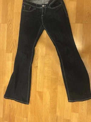 Mörkblå bootcut jeans med kontrastsömmar - Snygga mörkblå bootcut jeans med vita kontrastsömmar och klassisk femficksmodell. Jeansen har låg midja och vida ben, perfekta för en retroinspirerad look. Tillverkade i denim med en cool tvätt och detaljerade bakfickor.