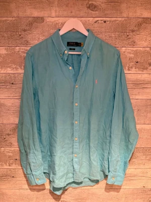 Ljusblå skjorta från Polo Ralph Lauren - Klassisk ljusblå skjorta från Polo Ralph Lauren med button-down krage och broderad logga på bröstet. Skjortan har långa ärmar, knappar framtill och är tillverkad i bomull. Perfekt för en clean och stilren look.