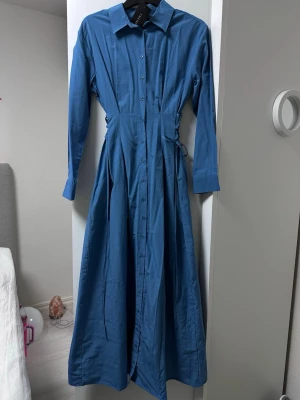 Blå skjortklänning med snörning - Snygg blå långklänning från Trendyol med klassisk skjortkrage, knappar hela vägen framtill och långa ärmar. Klänningen har snörningsdetaljer i midjan för en justerbar passform och är tillverkad i ett mjukt bomullstyg.