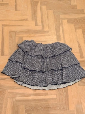 Rutig volangkjol med blå detaljer - Supersöt kort kjol med flera volanger och rutigt mönster i blått och vitt. Kjolen har elastisk midja och är perfekt för dig som gillar en lekfull och trendig stil. Passar till många olika outfits och ger en cool touch.