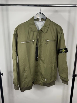 Olivgrön bomberjacka Stone Island - Snygg olivgrön bomberjacka från Stone Island med klassisk patch på ärmen. Jackan har två bröstfickor med dragkedja, två sidofickor och ribbade muddar. Perfekt för dig som gillar streetwear och vill ha en stilren look.