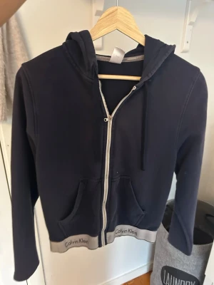 Mörkblå hoodie från Calvin Klein - Snygg mörkblå hoodie från Calvin Klein med dragkedja framtill, huva med dragsko och två fickor. Grå ribbade muddar med Calvin Klein-logga längs nederkanten ger en clean look. Perfekt för chill dagar eller när du vill ha en enkel men stilren outfit.