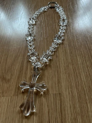 Kedjehalsband med stor korsberlock - Unikt halsband med transparant kedja och en stor korsformad berlock i klar plast. Materialet är en glasliknande plast. Pris går att diskutera