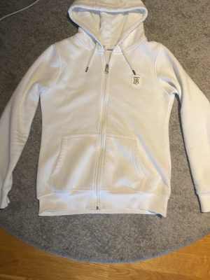 Vit hoodie med dragkedja från Burberry - Snygg vit hoodie från Burberry med hel dragkedja, huva med snörning och kängurufickor. På bröstet sitter ett elegant Burberry-monogram i guld. Perfekt för en clean och stilren look.