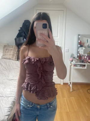 Ljuslila volang linnetopp - Säljer en ljuslila linnetopp med volangdetaljer och smala axelband. Toppen är croppad och har en rynkad design som ger en snygg och trendig look. Perfekt för dig som vill ha något unikt och stilrent.