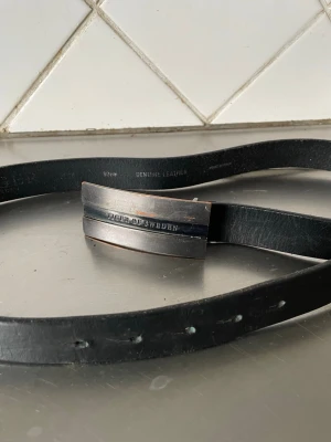 Svart skärp från Tiger of Sweden - Stilrent svart skärp från Tiger of Sweden med en metallfärgad, rektangulär spänne där loggan är ingraverad. Skärpet är gjort i äkta skinn och har en klassisk, minimalistisk design som passar till många olika outfits.