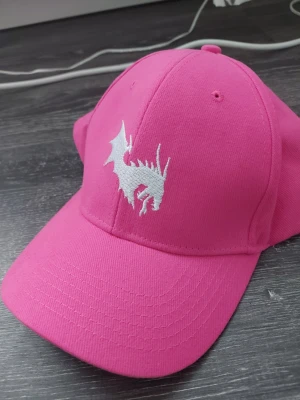 Rosa keps med drakbrodyr - Snygg rosa keps med böjd skärm och vit broderad drake framtill. Justerbar baktill för perfekt passform. Tillverkad i mjukt och slitstarkt tyg, perfekt för att lyfta din streetwear-look.
